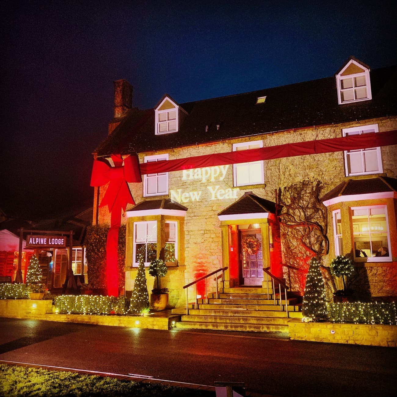 A cotswold house it adorned with a giant red bow, pea lights and a gobo showing happy new year. The   Dormy House hotel flood lit. 