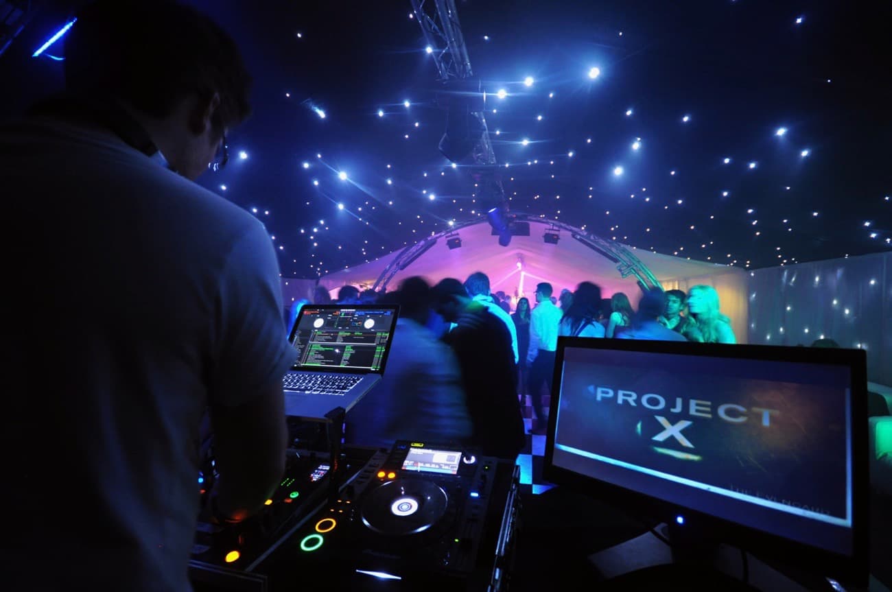 Dj stands at the decks whilst people dance in a marquee under a nightclub lighting rig