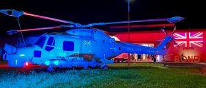 IMG_9855 A helicopter uplit in blue is in front of RNAS Yeovilton which is uplit in red for a party. The exterior of the building has a Union Jack flag projected onto it.