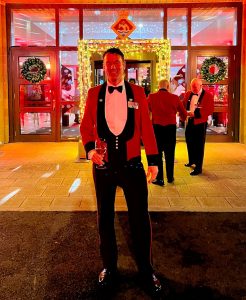 IMG_9864 The exterior of Royal Naval Air Station Yeovilton with James Bond themed lighting projected onto the front of the building which is uplit in red for a party. A man in military smart dress smiles at the camera. Behind him there is a truss archway with fairylight around it to enter the building.