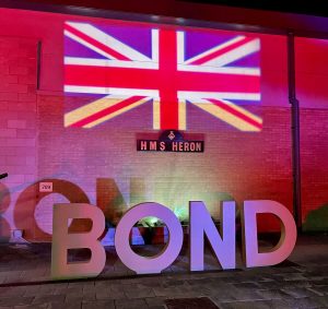 IMG_9871 A union jack flag is projected onto the exterior of a building which is uplit in red. Large freestanding letters spell out "Bond" in front of it.