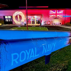 IMG_9908 The exterior of Royal Naval Air Station Yeovilton with James Bond themed lighting projected onto the front of the building which is uplit in red. There is a truss archway with fairylight around it to enter the building. A part of a helicopter is at the front of the image with "Royal Navy" on.