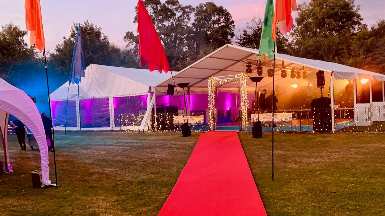 Two frame marquees on a tennis court ready for a summer party. A red carpet leads up to the front of the marquee