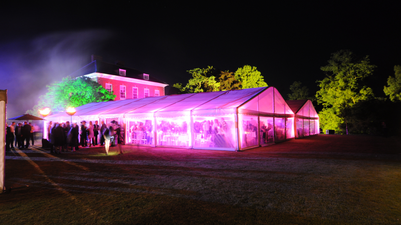 Two marquees at night on a grass tennis court outside a large country house. The marquees are beautifully lit with pink uplights