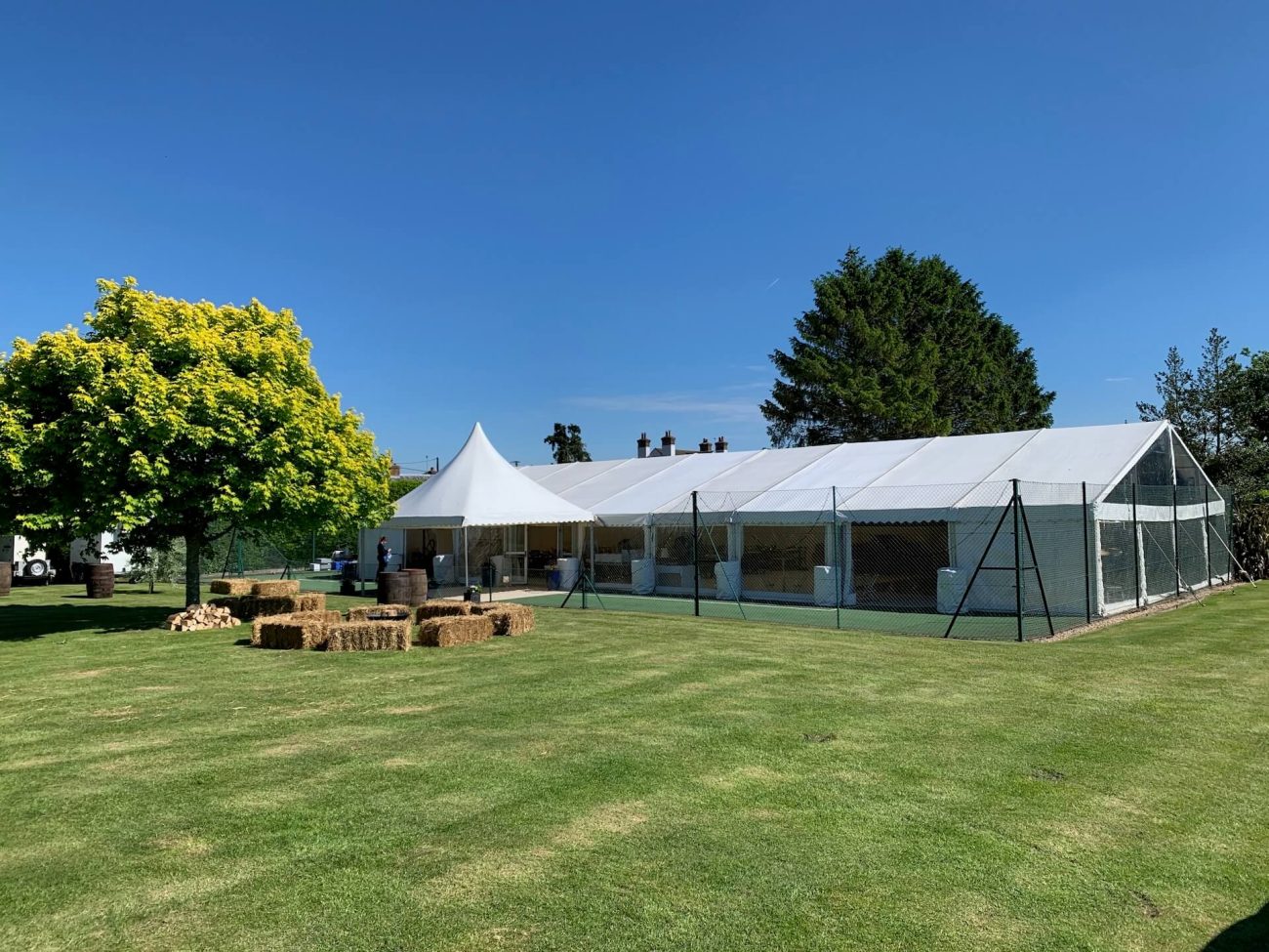 Marquee sits on a tennis court in the sunshine of a beautiful day. Hay bales sit in a circle around a fire pit
